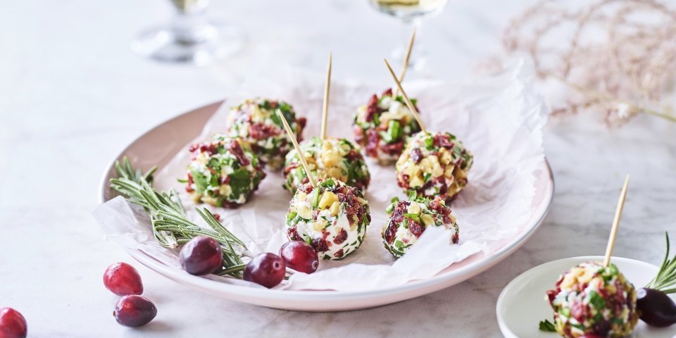 Kruidige kaasballetjes liggen op een licht roos bordje. Op de achtergrond staan 2 glaasjes aperitief met enkele veenbessen ter versiering. 