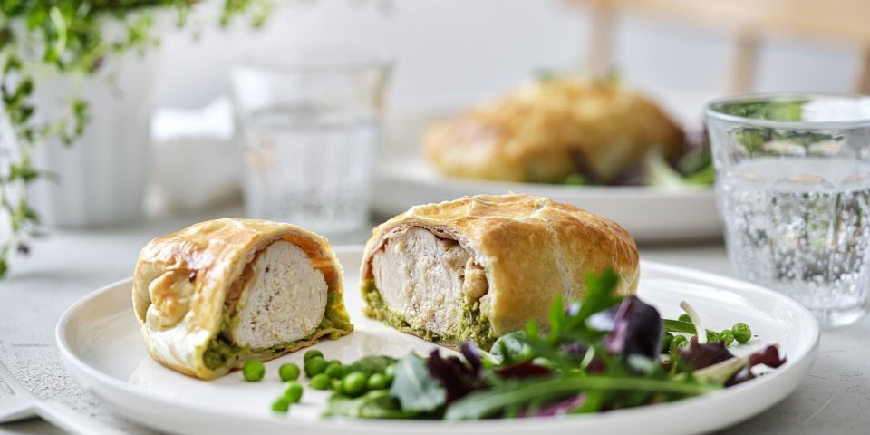 Kip wellington met gedroogde ham en erwtjespuree met munt Een mini wellington van kipfilet ligt opengesneden op een wit bord. Hierin zie je mooi een laagje erwtjespuree onder het witte vlees in het bladerdeeg pakje. Er is nog wat gemengde sla en erwtjes toegevoegd op het bord.