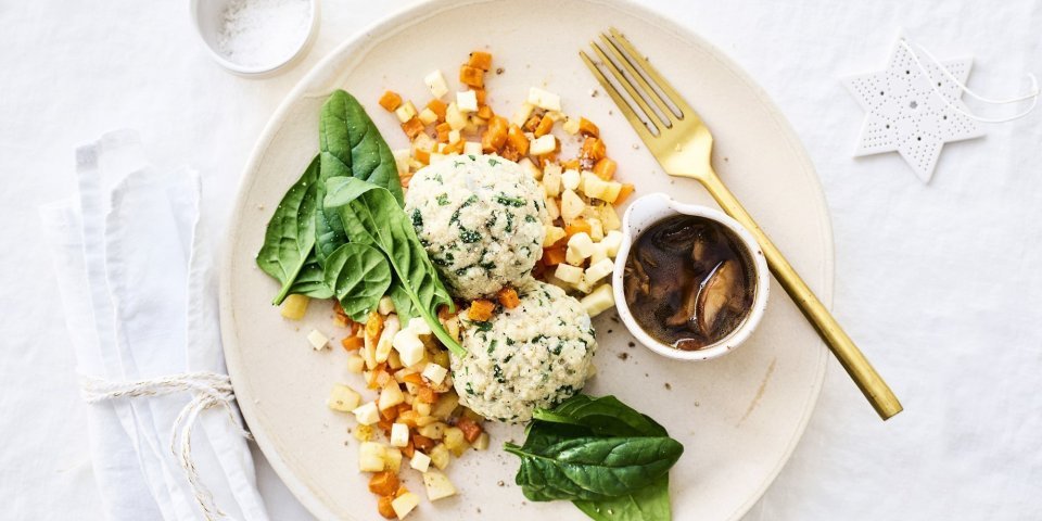 Knoedels, gemaakt van brooddeeg, liggen bovenop gestoofde blokjes peterseliewortel en zoete aardappel. Deze vegetarische ballen worden opgediend met een sausje van bospaddenstoelen. Het sausje staat mee op het bord in een potje. Goud bestek en kerststerren liggen langs het bord.