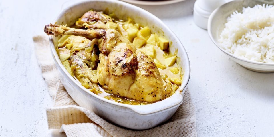 Konijn uit de oven met appel en curry Op tafel staat een witte ovale ovenschotel met hierin de gele currysaus met stukjes appel en ananas. Hier bovenop liggen de bouten te blinken. De ovenschotel staat op een beige linnen handdoek met rijst, borden en een witte pepermolen langs.