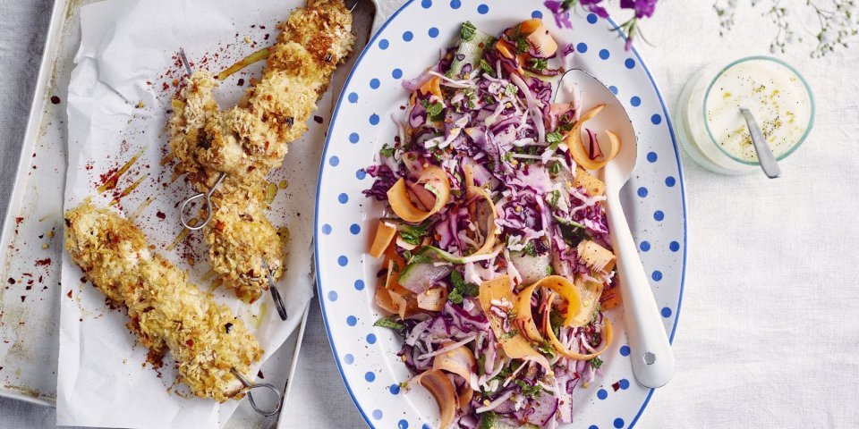 Krokante kipbrochette met een slaatje van kool, komkommer en wortel Krokante kipbrochette ligt nog de sidderen op een ovenplaatje. Daarnaast staat een kleurrijk groot bord met een salade op basis van rode kool, wortel en komkommer. De dressing op basis van yoghurt staat langs de salade.