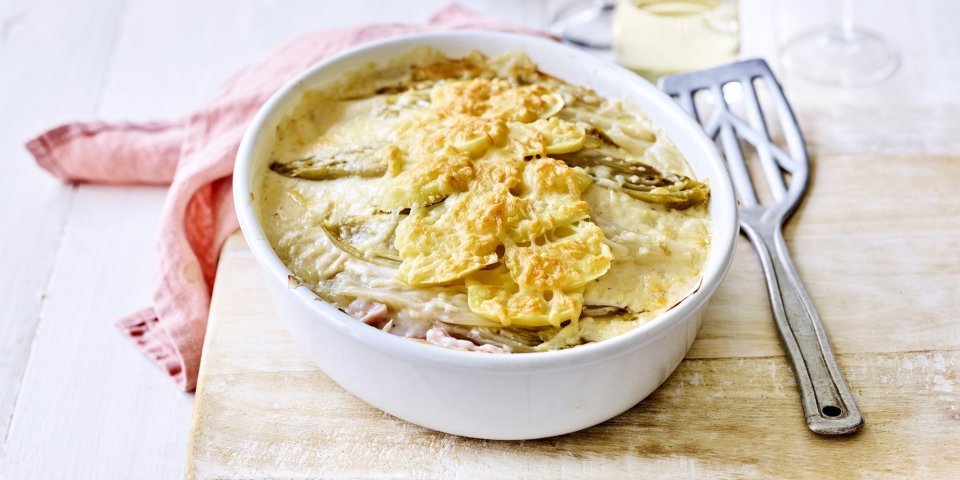 Lasagne van witloof met ham, kaas en aardappel Er staat een ovenschotel op een houten plank, op de tafel. Er ligt een lepel naar de ovenschotel die gevuld is met een lasagne van witloof, ham, kaas en aardappelen. Er ligt ook een coraalroze servette bij.