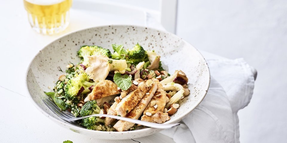 Een rijkgevulde quinoasalade boordevol verse groenten en kruiden zoals broccoli, paddenstoelen, rode ui, peterselie, koriander en munt. Bovenop de quinoa werd het varkensvlees geplaatst in schijfjes. Er staat een witbier bovenaan het bord, op de tafel.