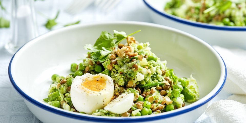 Een wit emaille bordje met blauwe rand is gevuld met de linzensalade. Het is een salade rijk aan groene groenten. Het gerecht is afgewerkt met een gekookt eitje. 