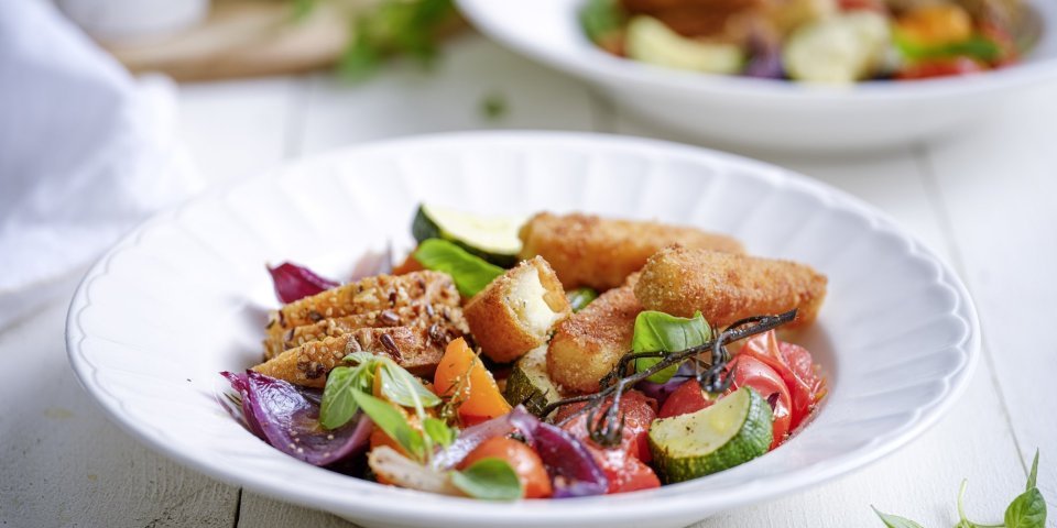 Maaltijdsalade met krokante berloumi Een heerlijk en rijkgevuld slaatje is geserveerd in een diep wit bord. De kleurtjes van de basilicum, tomaten, rode ui en courgette zijn uitgesproken en zien er fantastisch uit. Dit gerecht geeft je een boost.