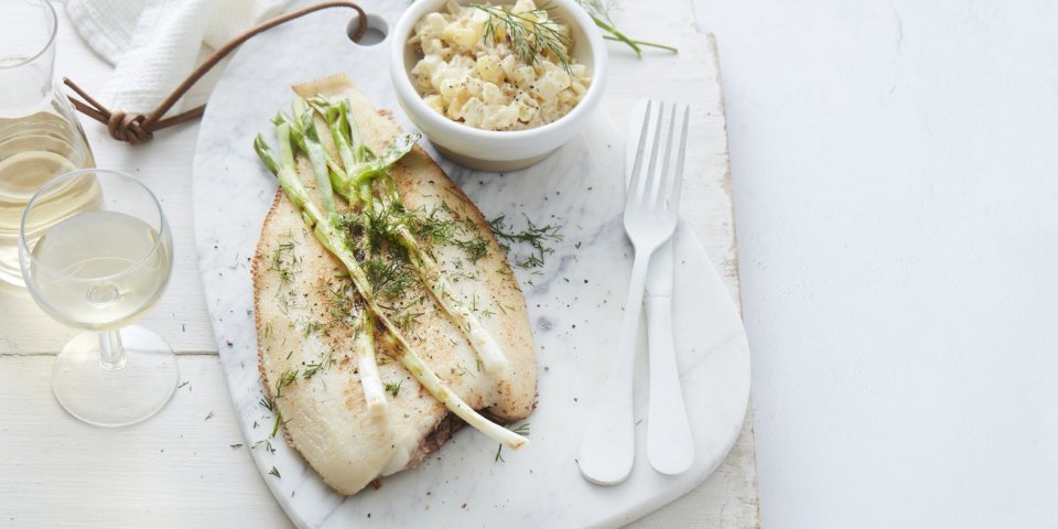 Macédoine van venkel en krieltjes met tongschar en gegrilde lente-ui De gebakken vis ligt op een stenen witte snijplank, waar de gigrilde lente-ui bovenop ligt. De Macédoine van venkel en krieltjes is opgediend in een apart wit potje. Er ligt wit bestek langs, met een glaasje witte wijn en een linnen servette. Het gerechtje is afgewerkt met verse dille.