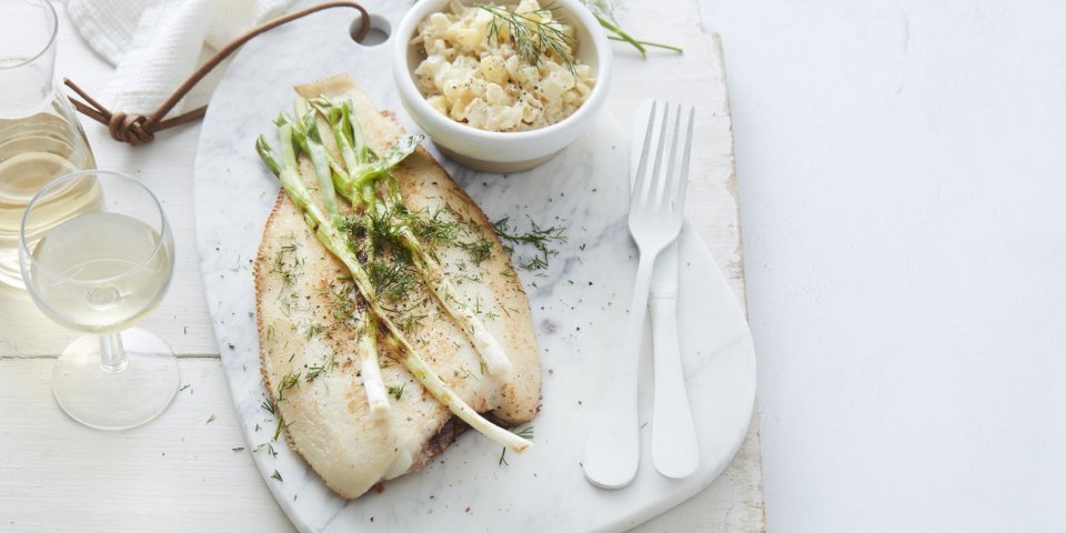 De gebakken vis ligt op een stenen witte snijplank, waar de gigrilde lente-ui bovenop ligt. De Macédoine van venkel en krieltjes is opgediend in een apart wit potje. Er ligt wit bestek langs, met een glaasje witte wijn en een linnen servette. Het gerechtje is afgewerkt met verse dille.