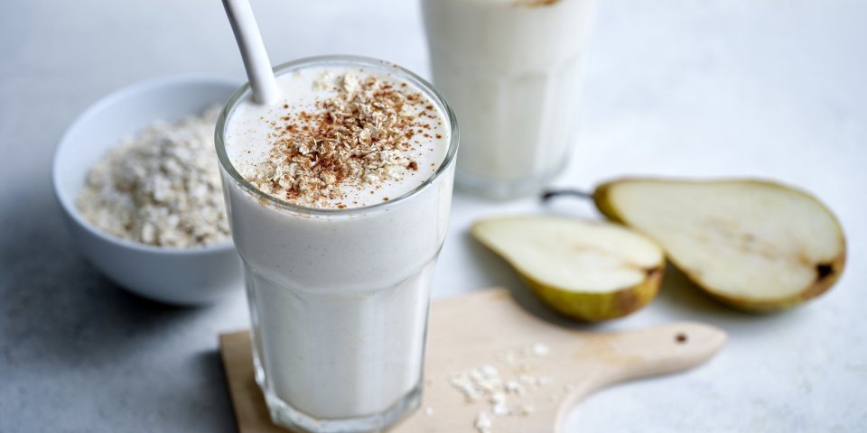 Milkshake met havermout en peer Twee klaargemaakte milkshakes staan op het lichtgrijze aanrecht te pronken. Er ligt een peer langs die doormidden gesneden is en wat havermoutvlokken gemorst. De shakes zijn afgewerkt met wat kaneelpoeder.