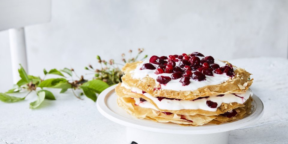Millefeuille met kersen Grote ronde millefeuille met kersen en verse roomkaas. Deze staat op een witte taartschaal op een bleke houten tafel. Op tafel ligt ook een takje groen voor extra sfeer.