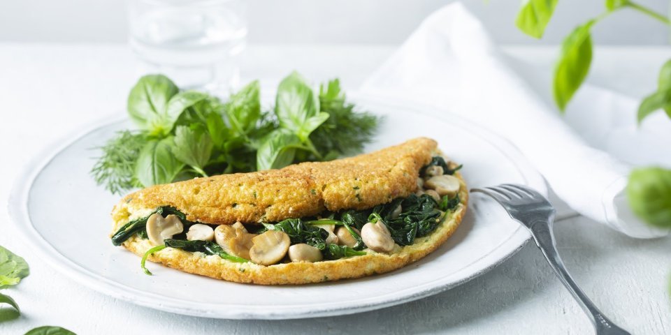 Omeletsoufflé met champignons, spinazie en kaas Een heerlijke dichtgevouwde omeletsoufflé gevuld met champignons, spinazie en kaas ligt op een wit bord. Het gerechtje werd afgewerkt met een slaatje van verse kruiden, denk maar aan basilicum en dille.