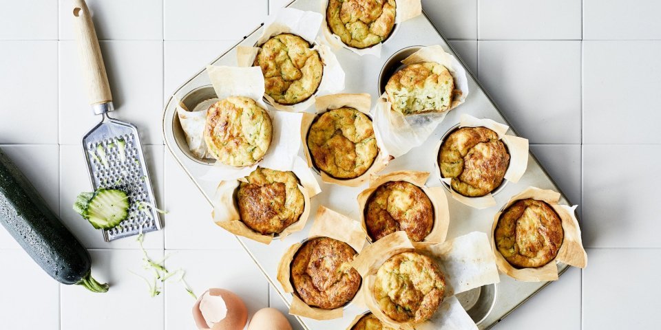 Ontbijtmuffins met courgette, yoghurt en Belgische kaas Ontbijtmuffins met courgette, yoghurt en Belgische kaas in een grote muffinvorm. Klaar om eruti te halen en op te eten. Naast de bakvorm ligt een verse courgette en lege eierschalen voor de sfeer.