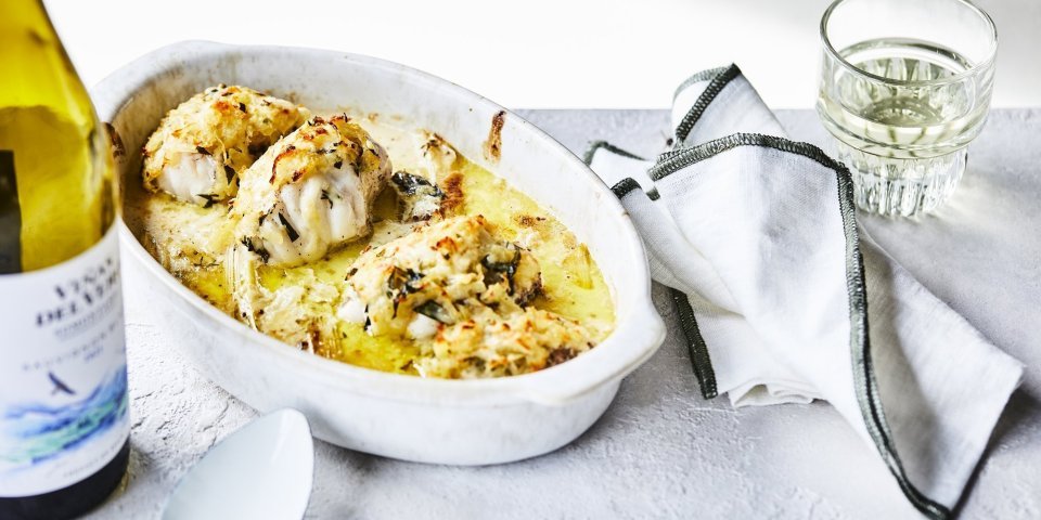 Ovenschotel met zeeduivel Een ovenschotel staat klaar op tafel om meteen te beginnen. De schotel is gevuld met stukken zeeduivel en prei in een mosterdsausje. Er ligt een krokant korstje op van geraspte aardappel en kaas. Er staat een flesje wijn langs en een witte scheplepel.