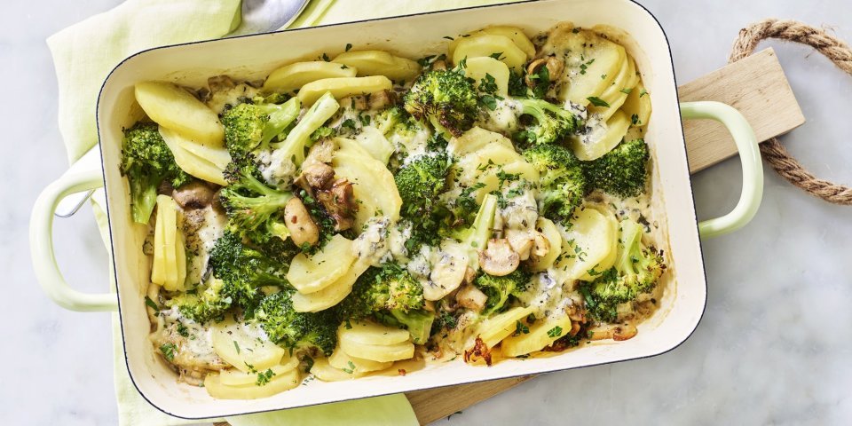 Ovenschotel van aardappel, broccoli, champignons en blauwe kaas Een ovenschotel van aardappel, broccoli, champignons en blauwe kaas staat op het midden van het marmere aanrecht op een houten snijplank. Verse kruiden zorgen voor een extra groene touch!
