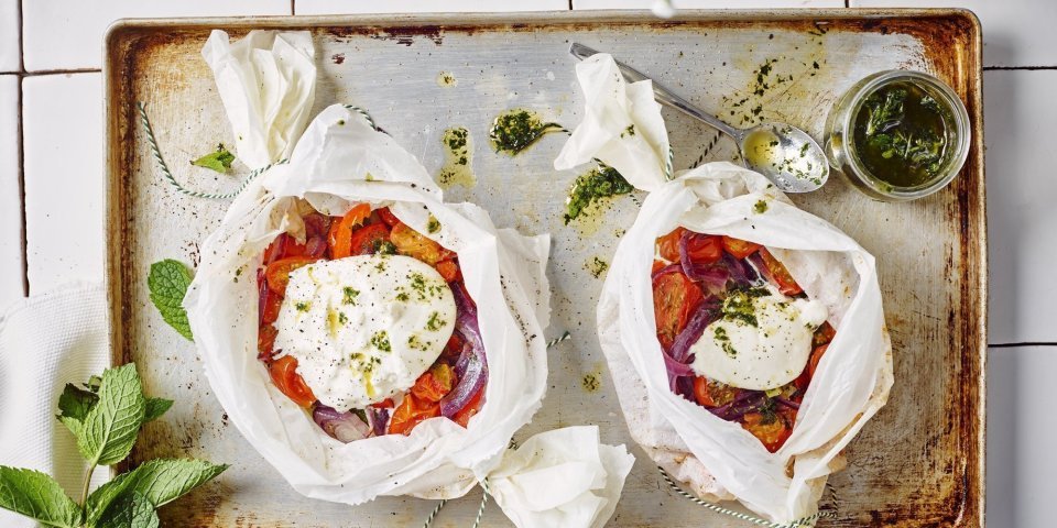 Twee papillotten liggen op een ovenplaat. De papillot is gemaakt in een wit boterpapiertje waarin de groenten een mooi contrast in geven. Denk maar aan de rode paprika's en tomaten. De burrata ligt er bovenop en de muntolie staat in een bokaaltje naast de papillotten.