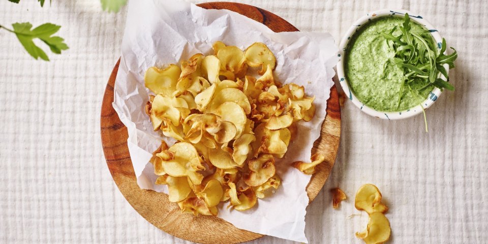 Op tafel ligt een kalkwit tafelkleed, met hierop een houten bordje met wit boterpapiertje en chips van pastinaak op. Er staat een dippotje langs met de dip van broccoli en roomkaas. 