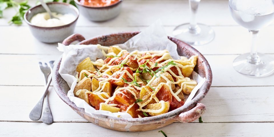 Heerlijke patatas bravas geserveerd in een groot diep bord, afgewerkt met een pittige tomatendip en aïoli. Twee gekoelde watertjes staan rechts van het bord. Klaar om de tapas tafel aan te vallen.