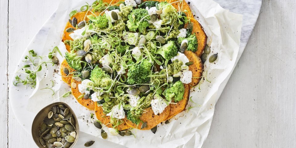 Pizza van zoete aardappel met geitenkaas en groene groenten Klaargemaakt pizza van zoete aardappelschijfjes ligt op een snijplank van marmer. De pizza werd belegd met geitenkaas en groene groenten.