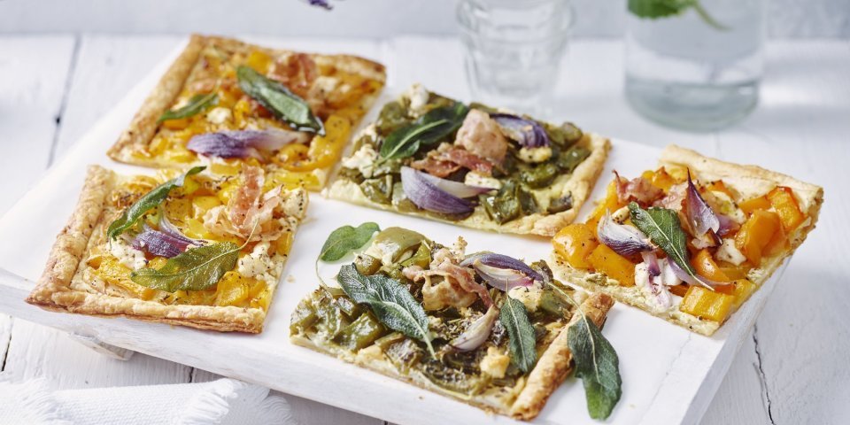 Plaattaart met paprika, spek en salie Plaattaart met paprika, spek en salie ligt aangesneden klaar op tafel. Er staat een fles water met glaasje langs. Paarse bloemen zorgen voor een extra toets in het beeld.