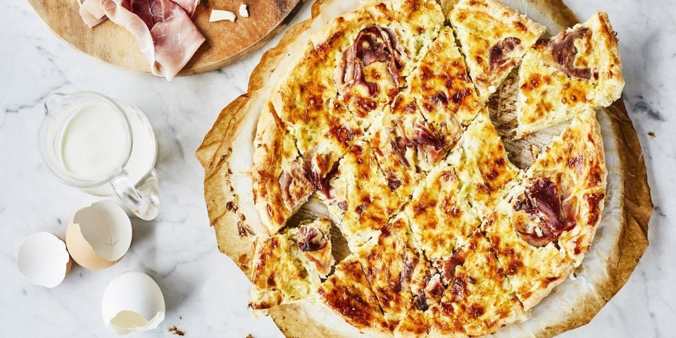 Quiche Lorraine Vers uit de oven staat deze quiche te schitteren op het marmere aanrecht. Hij is aangesneden in kleinere stukjes om aan te vallen tijdens het aperomoment. Er liggen enkele eitjes naast ter decoratie en verse boerenham.