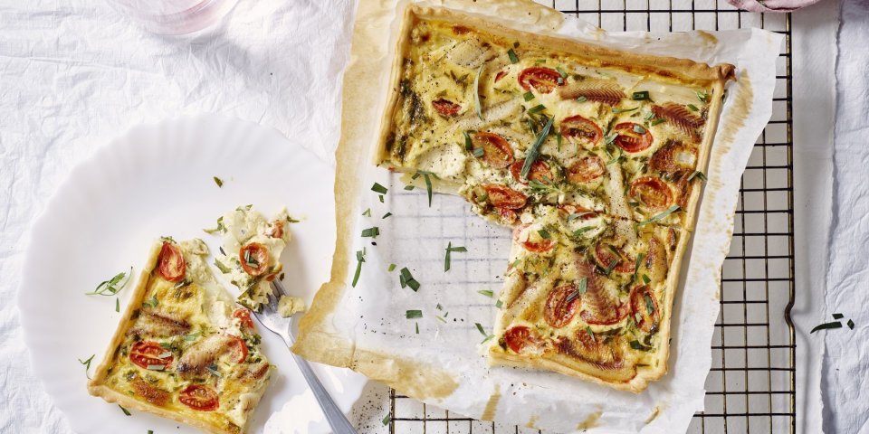 Vers gebakken uit de oven staat de quiche met asperges, tomaten en gerookte paling te blinken op tafel. Er werd al een stukje aangesneden en op een bordje gepresenteerd. Roze linnen servet en waterkaraf zorgen voor de sfeer in de foto. 