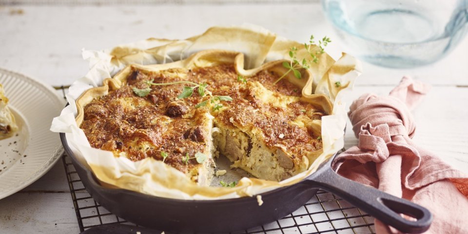 Quiche met bloemkool en worst Op het aanrecht ligt een zwarte rooster met hierop de quiche in een gietijzeren pan. Hierbij ligt een roze servet, mes en bordje waarop al een stukje quiche op ligt. De quiche is afgewerkt met enkele verse blaadjes oregano.