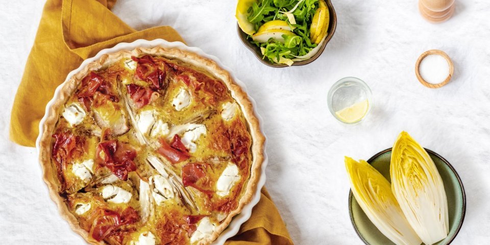Quiche met witloof, peer en gedroogde ham Een ronde quiche met wiltoof staat op een okkerkleurige handdoek op het aanrecht.