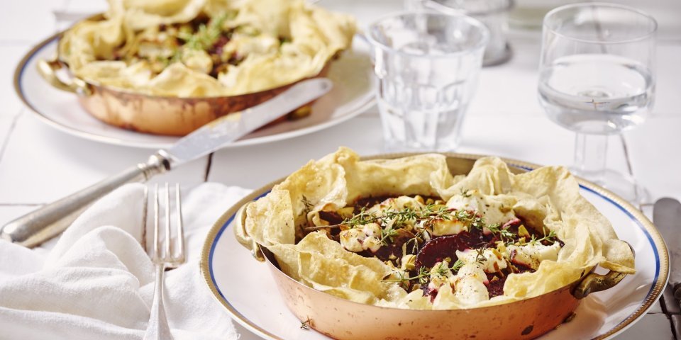 Aan tafel! Quiche van filodeeg met geitenkaas en rode biet staan klaar op tafel. De quiches zitten in een kopperen schaaltje en staan op een wit bord. Het bestek en de witte servette liggen langs het bord. Op tafel staan ook waterglazen en een glazen peper- & zoutvat.