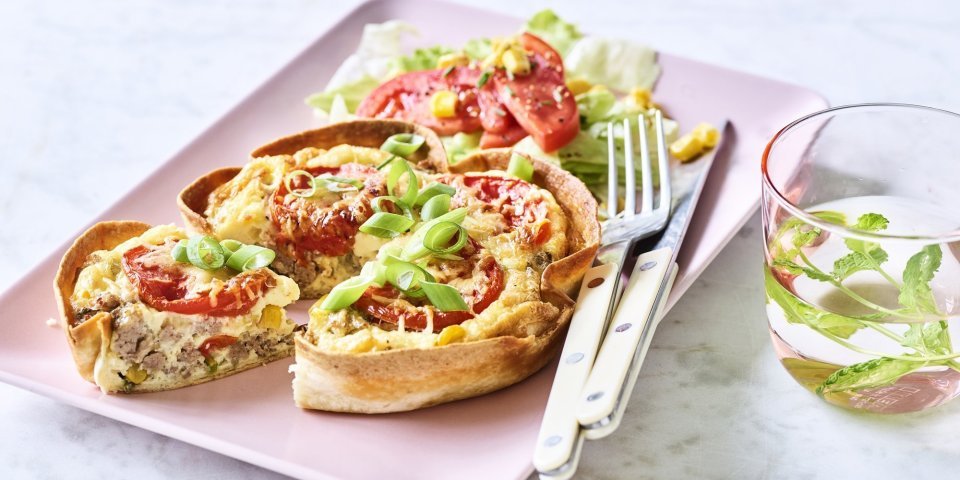 Quiche van tortilla met pittig gehakt, tomaat en mais Een klaargemaakte quiche ligt op een roze en rechthoekig bord klaar om te eten. De quichebodem bestaat uit een wrap met hierin het quichebeslag en de groenten.