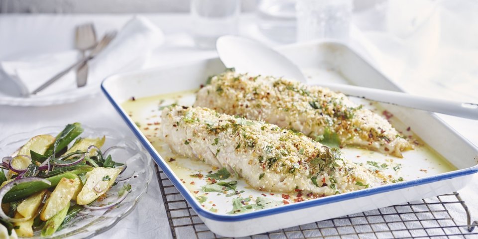 De gegrilde roodbaars uit de oven ligt op een witte bakplaat en staat op een rooster. De crumble zorgt voor een zomerse sfeer in het gerecht. Naast de ovenschaal staat een bordje met wat aardappelsalade uitgeschept. 