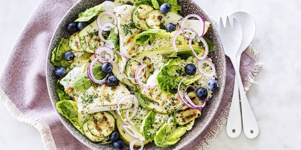 Een diep bord gevuld met sla, gegrilde zeetong, courgette, peer en blauwe bessen. Onder het bord ligt een paars servette en wit bestek om meteen dit gerechtje te verorberen. 