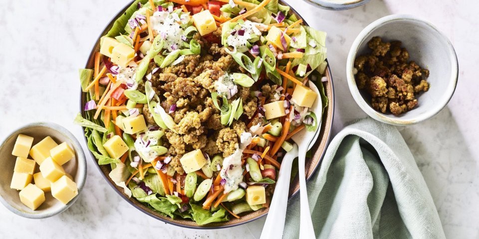Salade met gehakt en ranchdressing Een grote salade kom staat op het aanrecht, hij is gevuld met gemengde sla, komkommer, tomaten, worteltjes, stukjes kaas en gebakken gehakt. We werkte de salade af met een ranchdressing op basis van zure room en verse kruiden.