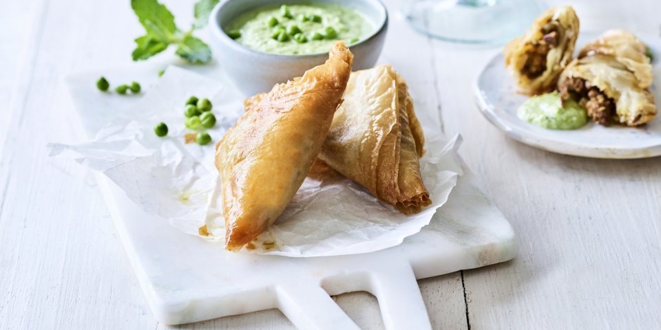 Klaargemaaakte samosa met lamsvlees liggen gepresenteerd op een marmer plankje. Ze worden opgediend met een frisgroene dip van munt en erwtjes.
