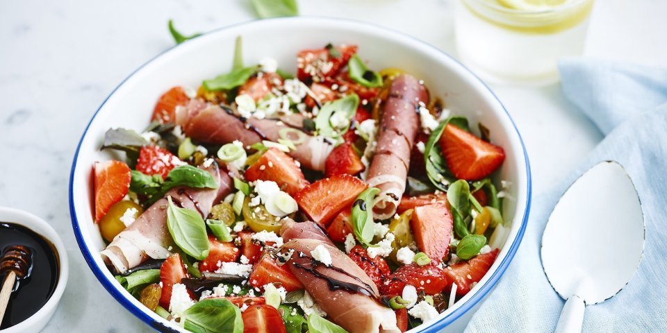 Rijkgevulde salade met aardbeien, geitenkaas en gandaham. Afgewerkt met balsamico. Er zijn twee frisse waters boven de kom met elk een citroentschijfje in. 