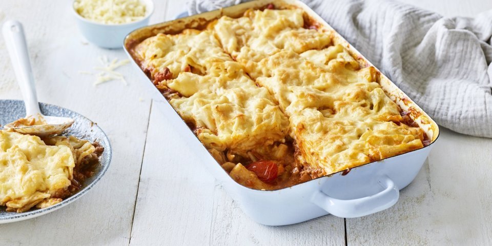 Slowcooked mac & cheese lasagne Een lichtblauwe ovenschotel staat op een houten witte tafel. De ovenschotel is gevuld met een mac & cheese én een tomatensaus, waardoor hij eerder weg heeft van een lasagne. Er is al 1 portie uitgeschept en wil al beginnen aan de volgende. Laat het niet koud worden.