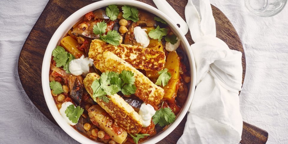 Stoofpot van aubergine en pompoen met Berloumi Op tafel ligt een wit tafelkleed met hierop een donker houte plank. Hierop staat het stoofpotje met een witte geknooptje handdoek langs. Er staat een glaasje langs met knoflookyoghurt en twee waterglazen. Het groentestoofpotje heeft een mooie warme kleur door de oranje pompoen, groene korianderblaadjes en de gegrilde kaas.
