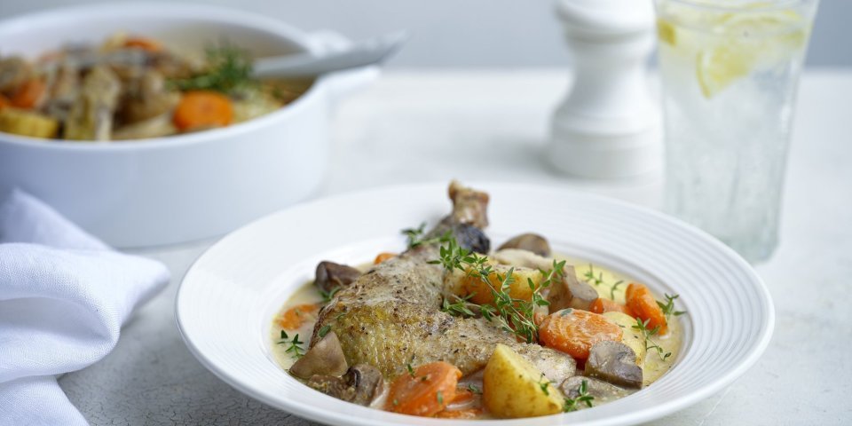 Een witte stoofpot staat op tafel. Deze keer is het eentje met kip, aardappelen en groentjes die gegaard zijn in melk. Er staat ook een glas water langs met een schijfje citroen. 