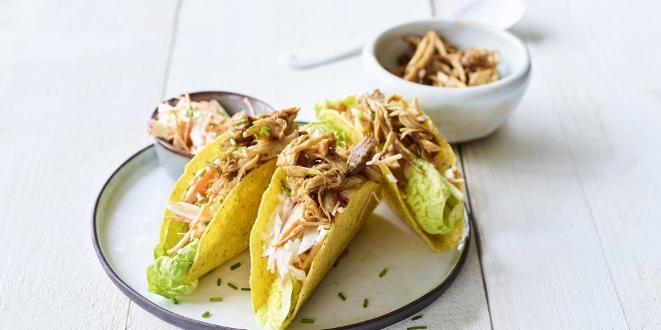Taco met traaggegaarde varkensspiering en een koolsalade Krokante taco's liggen op een bordje, ze zijn gevuld met stukjes varkensvlees en een slaatje van kool.