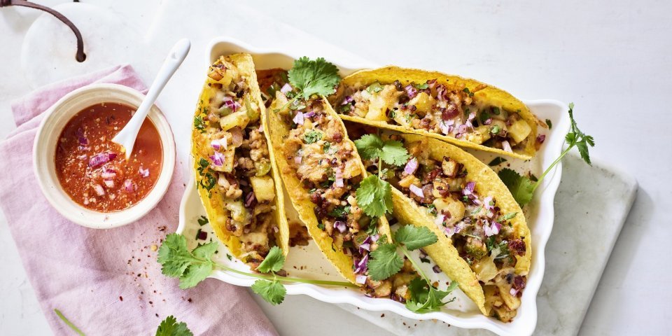 Vier tacos liggen klaar in een ovenschaal om aan te vallen. De pittige tomatendip staat links van de schotel op een roze servette. Het gerechtje is afgewerkt met verse korianderblaadjes.