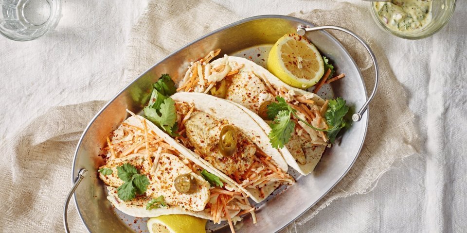 Taco’s met pladijs, koolsla en tartaarsaus Drie wraps liggen gepresenteerd in een ovenschotel van inox. Deze staat op een gedekte tafel met wit linnen kleed. Er liggen twee citroenhelften bij de tortilla's en verse koriander.