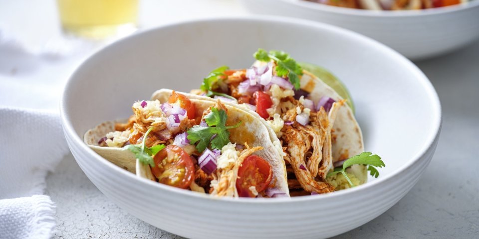 In een diep wit bord liggen twee gevulde taco's met pulled chicken, afgewerkt met tomaat, rode ui en geraspte Passendael kaas. Om wat extra kleur te geven liggen er enkele blaadjes koriander op. Wij serveren ze met frisgekoeld biertje, smakelijk!