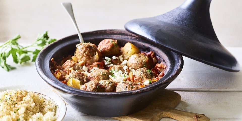 Tajine met gehaktballetjes van lamsvlees Zwartkleurige tajine met gehaktballetjes van lamsvlees, afgewerkt met verbrokkelde feta. Er staat een portie couscous naast de tajine.
