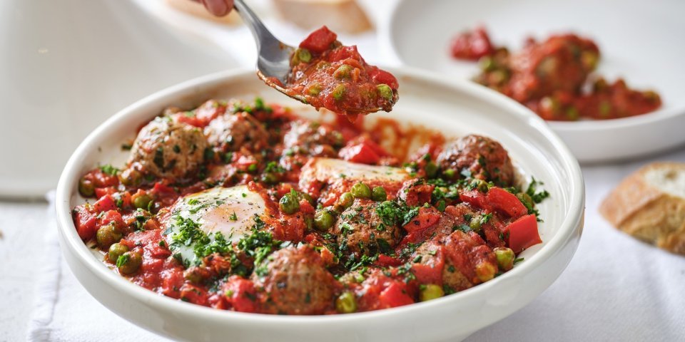 Tajine met gehaktballetjes, tomatensaus en ei Op tafel staat een witte tajine heerlijk gevuld met gehaktballen in een rijke tomatensaus met eitjes. Er wordt net een portie uitgeschept.