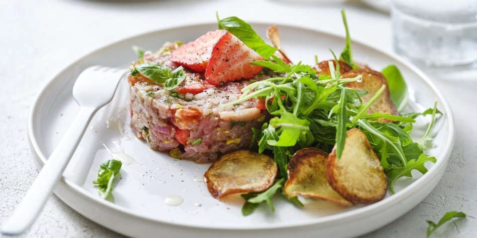 Tartaar van kalfsvlees met aardbei en aardappelplakjes Zoek je een alternatief voor steak-tartaar? Wel dit is een zomerversie, nl eentje met kalfsvlees, aardbeien, tomaatjes en verse kruiden. We werkten het gerechtje af met gebakken aardappelplakjes en verse rucola. Er ligt een wit vork en servette naast om meteen aan te vallen.