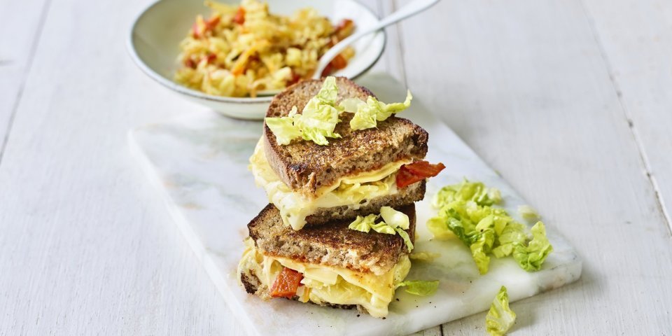 Toast met kazen van bij ons, kimchi en chilisaus Een heerlijke cheesy toast ligt op een marmere snijplank. De toast is gevuld met 2 Belgische kazen, kimchi en chilisaus.