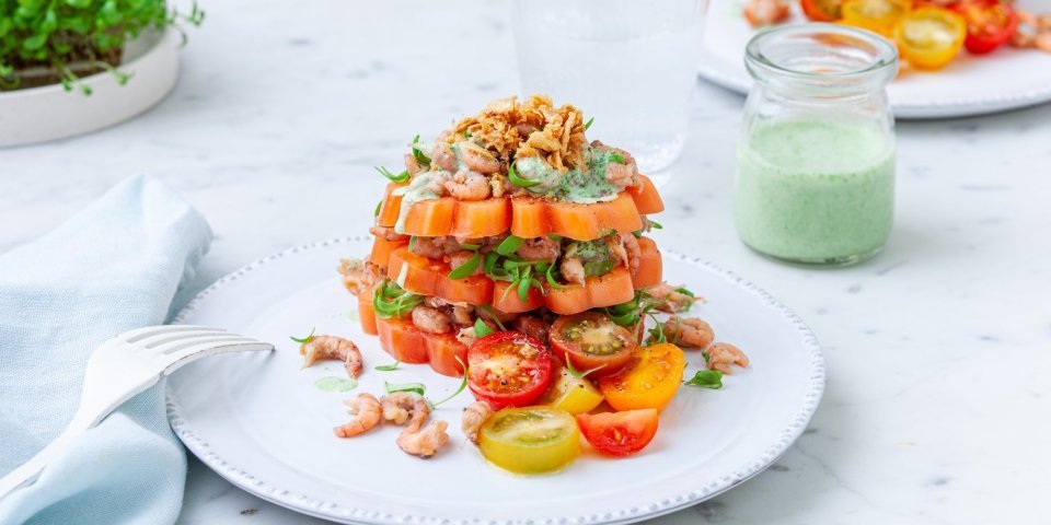 Tomaattorentje met grijze garnalen Een bord met daarop drie laagjes gesneden coeur de boeuf. Daarachter staat nog een bord met een potje met dressing en tuinkers.
