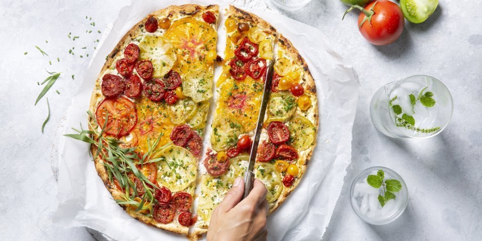 Tomatentaart met roomkaas en verse kruiden Een kleurrijke tomatentaart staat op tafel op een wit boterpapiertje. Ze wordt aangesneden door een onbekende vrouw met een koksmes. Rechtsboven liggen nog enkele tomaten om ze extra in de picture te zetten.