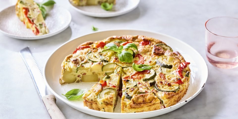 Een heerlijke aardappeltortilla ligt aangesneden op een groot wit bord. Er werden al twee stukjes uitgesneden en liggen geserveerd op kleine bordjes en afgewerkt met basilicum. Er staan licht gekleurd roze glazen langs het gerecht om de foto nog meer kleur te geven.