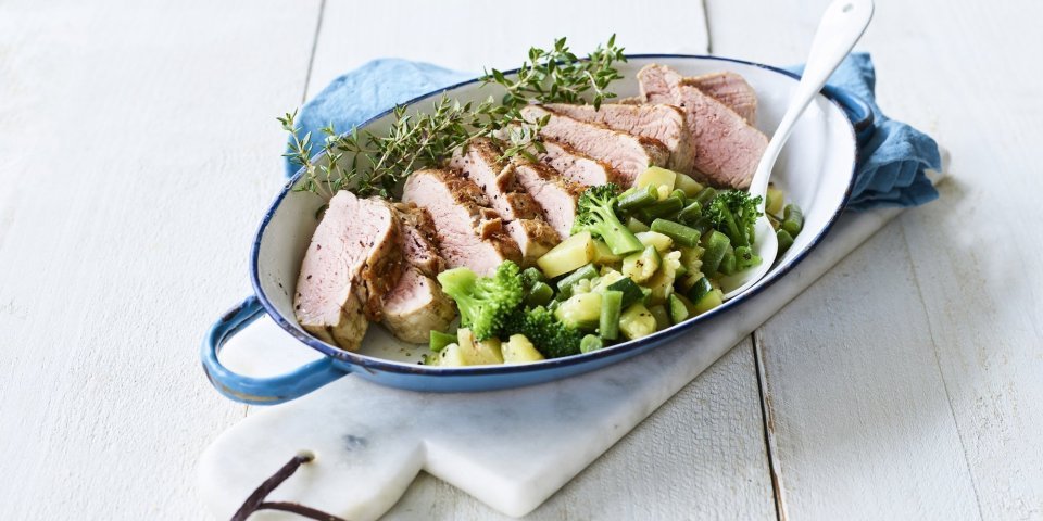 Varkenshaasje in de oven met macedoine van groene groenten Een heerlijk varkenshaasje uit de oven ligt in een blauwe ovenschaal, vergezeld met groene groenten en verse tijm.