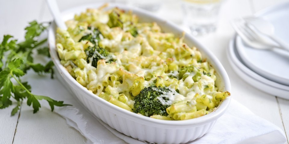 Vegetarische Mac'n cheese met witte bloemkoolsaus en broccoli Een grote ovenschaal staat op een houte witte tafel. Hij staat op een wit lapje stof. De ovenschotel is gevuld met macaroni, broccolie en witte bloemkoolsaus. Er staan enkele bordjes langs met wit bestek om direct aan te vallen.
