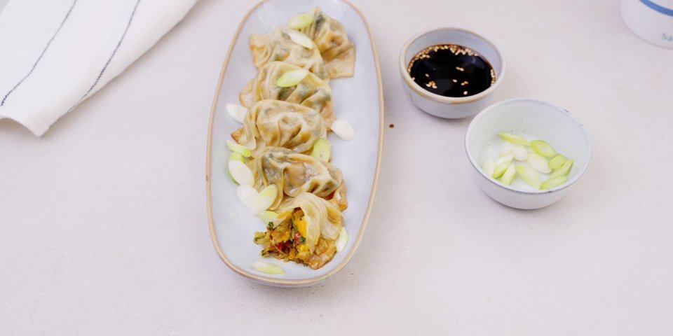 Vegetarische dim sum Vijf dim sum's liggen op een langwerpig aperobordje. Er is eentje doormidden gebeten, waarin je de heerlijke mix van groenten en kruiden ziet. Er staat een potje met sojasaus langs en enkele versgesneden pijpajuintjes. Hap maar toe!